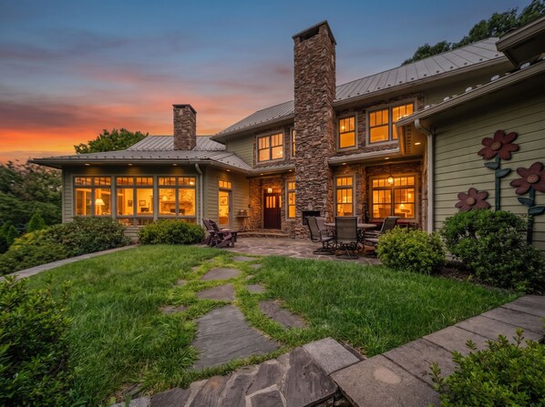 A two-story house with a stone chimney, large windows, and a stone pathway leading to a patio with outdoor furniture.