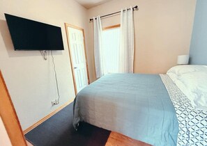A bedroom with a bed, a flat-screen TV mounted on the wall, a door, and a window with curtains. - Foosball|Cottage by McKennan|Fenced inYard (Sioux Falls)