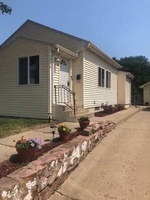 A single-story mobile home with a front porch, a small front yard with potted plants, and a stone retaining wall. - Foosball|Cottage by McKennan|Fenced inYard (Sioux Falls)