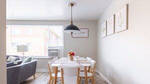A dining area with a white table, white chairs, a black pendant light, and a window with blinds.