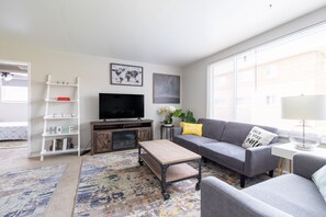 A modern living room with a flat-screen TV, grey sofa, coffee table, and a bookshelf.