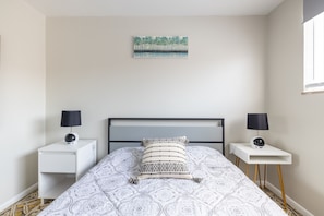 A bedroom with a bed, two bedside tables, a nightstand, a wall-mounted painting, and a window.
