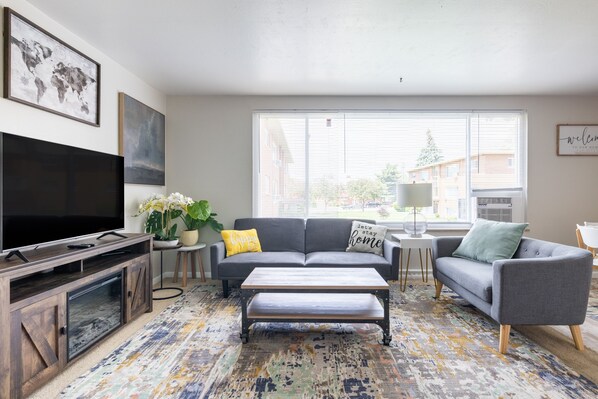 A modern living room with a flat-screen TV, grey sofas, a coffee table, and a patterned rug.