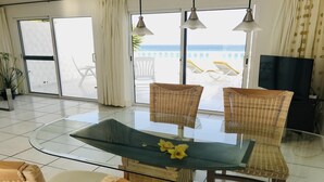 A modern living room with a glass table, wicker chairs, a TV, and a view of the sea through large windows.