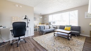 A modern living room with a black office chair, a gray sofa with a yellow pillow, a patterned rug, and a dining area with a white table and chairs.