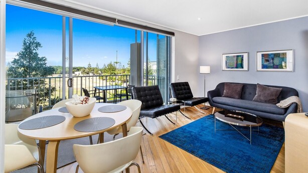 A modern living room with a balcony, a sofa, a coffee table, and a dining area with chairs and a table. - PEP 4305/6 - 2 Bedroom Apartment (Kingscliff)