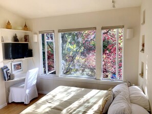 A bedroom with a bed, a TV on a shelf, and three windows with autumn foliage views. - Serene Montclair Getaway (Oakland)