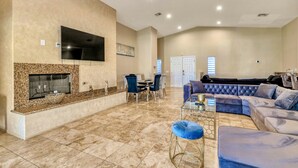 A spacious living area with a fireplace, a large TV mounted on the wall, a dining table with chairs, and a blue sectional sofa. - Private House with Pool and Spa (Desert Hot Springs)