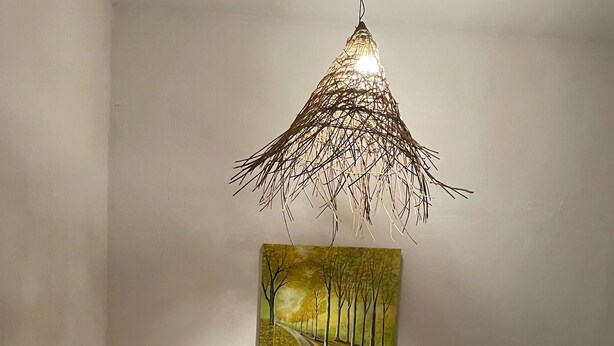 Une pièce avec un fauteuil vert, une petite table d’appoint en bois sur laquelle est posée une plante, un tableau représentant une route bordée d’arbres et un luminaire suspendu.