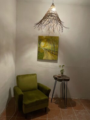 Ein Zimmer mit einem grünen Sessel, einem kleinen Holztisch mit einer Pflanze, einem Bild einer von Bäumen gesäumten Straße und einer Pendelleuchte.