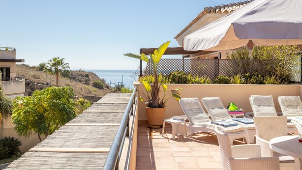 Un balcon avec vue sur la mer, des fauteuils de jardin blancs et une plante en pot.