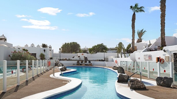A swimming pool with a curved design, surrounded by white buildings and palm trees.