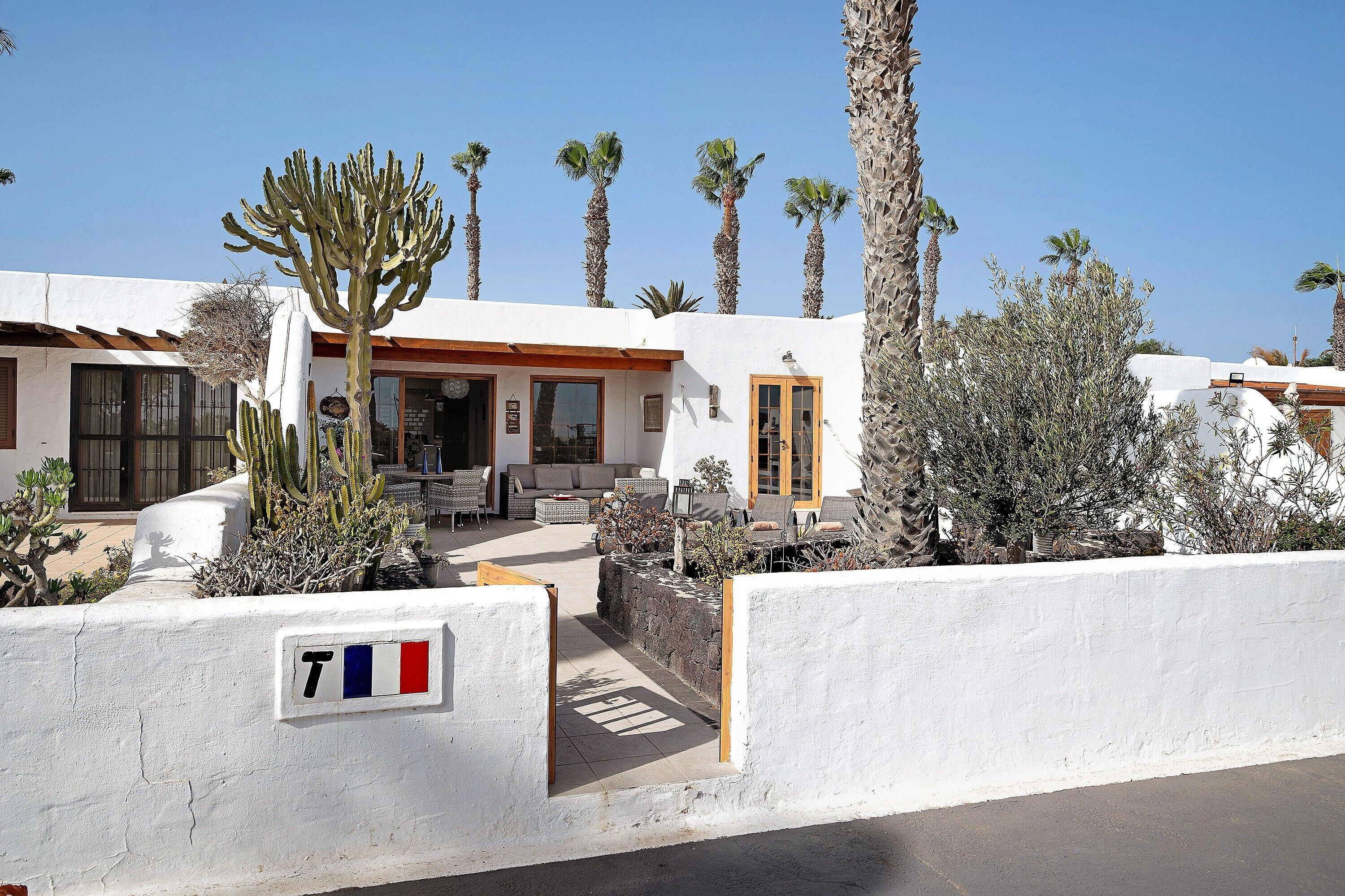 Un bâtiment blanc avec un panneau en forme de T, des palmiers et des tables à l’extérieur.