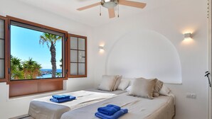 Un dormitorio con dos camas, un ventanal con vistas a palmeras y al mar, un ventilador de techo y luces empotradas.