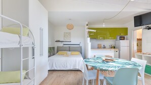 A dorm-style room with bunk beds, a single bed, a dining table with chairs, a kitchen area with green cabinets, and a TV mounted on the wall.