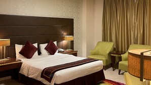 Une chambre d’hôtel avec un grand lit, deux tables de chevet avec des lampes, un fauteuil vert, une petite table ronde et un tapis coloré.
