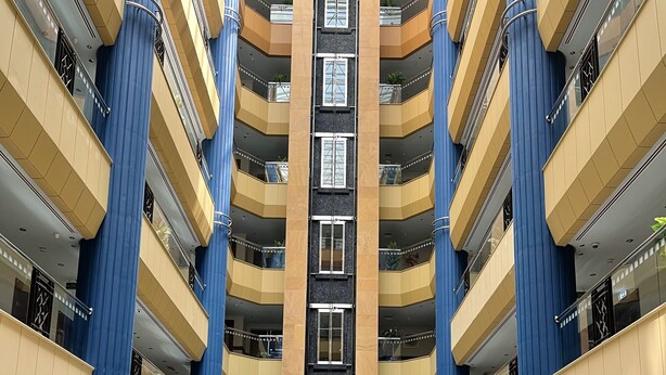 Un atrium à plusieurs niveaux, doté d’un escalier central, de colonnes bleues et beiges, et d’un espace détente avec des canapés et une table basse.