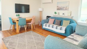 A living room with a blue sofa, a wooden dining table with chairs, a TV mounted on the wall, and a window with a view. - Lake, mountain view cottage - Entire house (Rotorua)