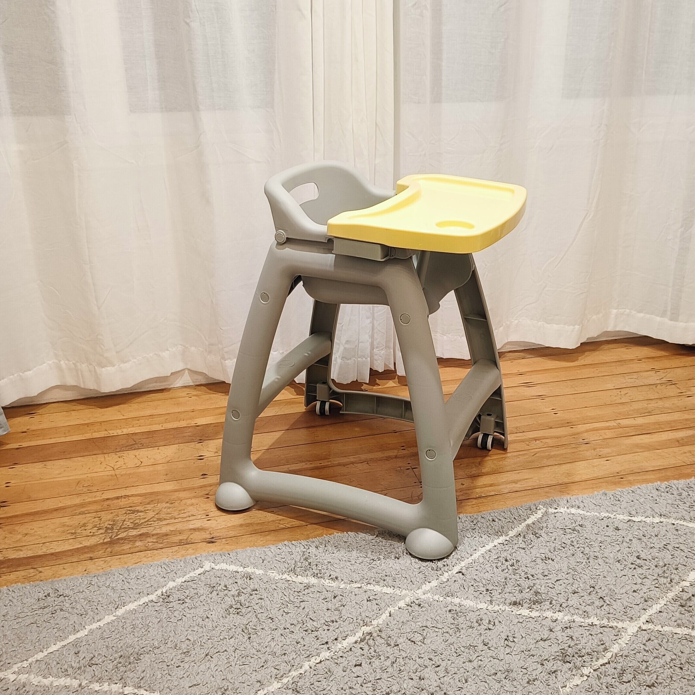 A grey and yellow high chair with wheels, placed on a wooden floor with a grey rug.