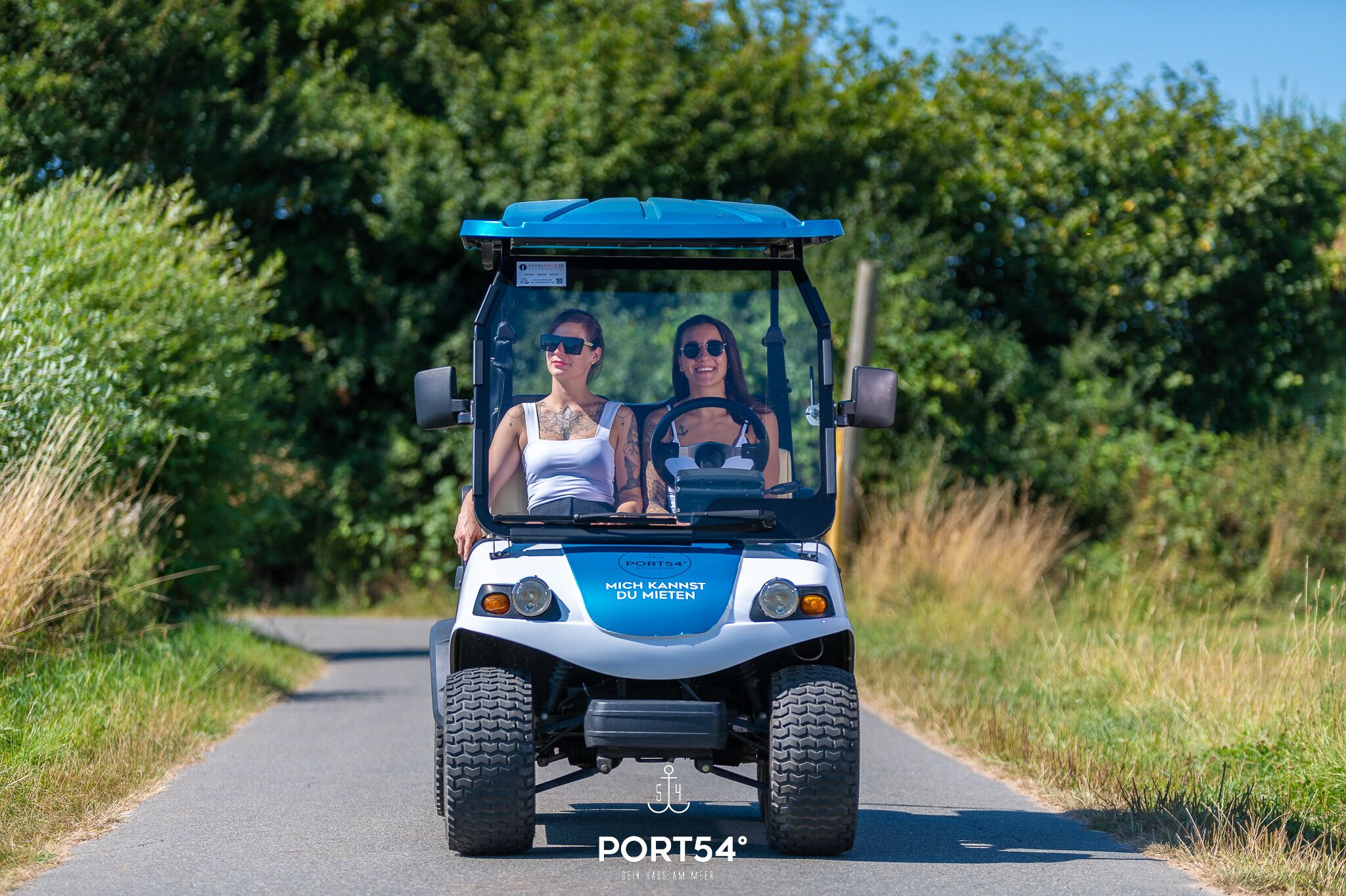 A golf cart with two people on a paved road surrounded by greenery.