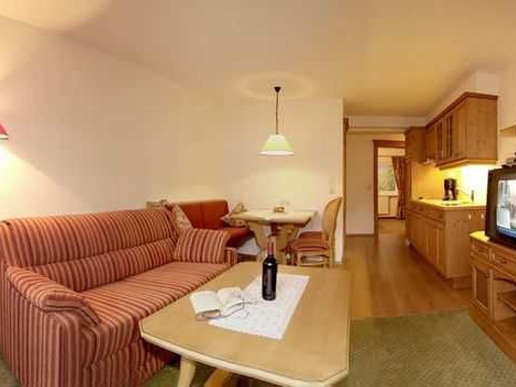 Un salon avec un canapé, une table sur laquelle se trouvent une bouteille et un livre, un téléviseur, et un coin cuisine à l’arrière-plan.