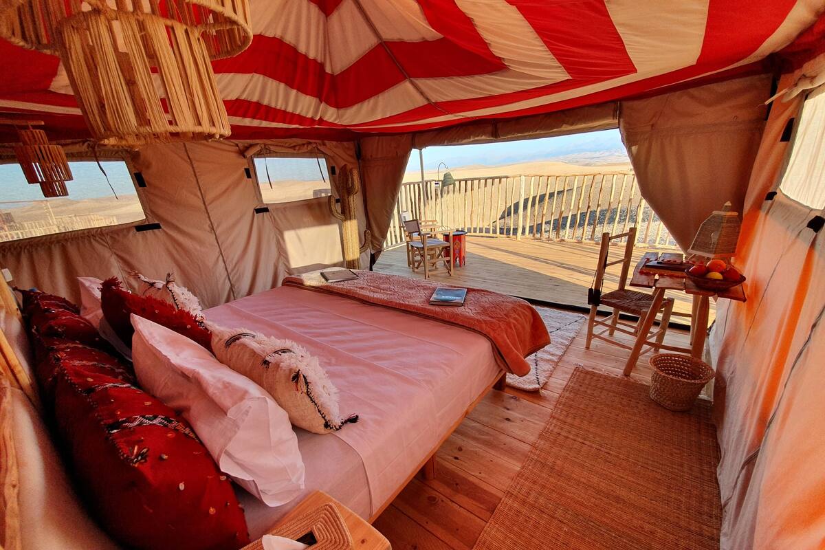 A tented accommodation with a bed, pillows, a table with a bowl of fruit, and a view of the desert.