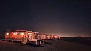 Filas de cabañas con cubierta de madera y techo rojo, ubicadas sobre terreno arenoso bajo un cielo oscuro.