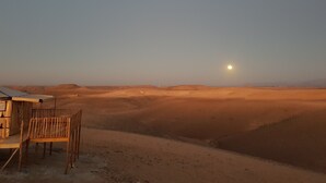 Paisaje desértico con una tienda de campaña y una silla de madera en primer plano, y una luna brillante en el cielo.