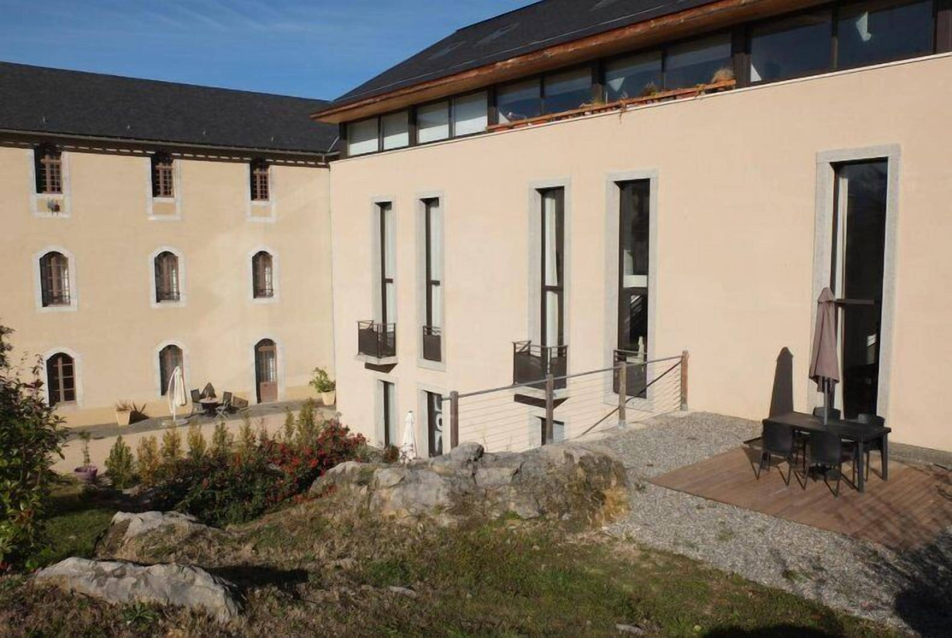 Un edificio con múltiples ventanas y balcones, un patio con mobiliario exterior y un muro de piedra al frente.