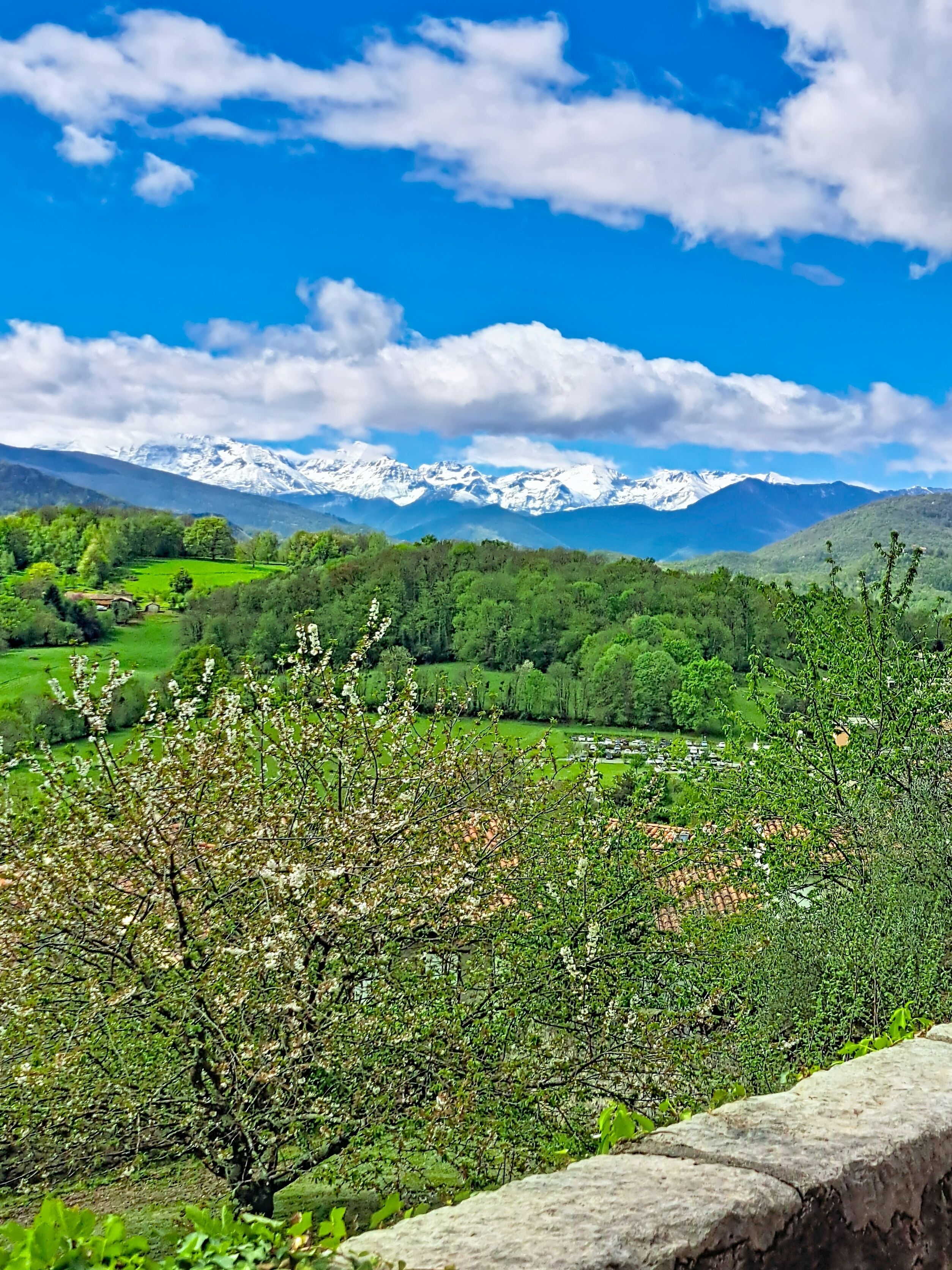 A mountainous landscape with snow-capped peaks, green valleys, and a stone wall in the foreground.