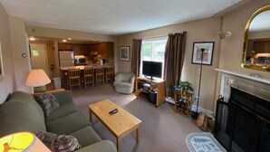 A living room with a sofa, coffee table, TV, and fireplace. There is a kitchen in the background.
