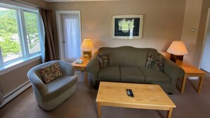 A living room with a sofa, armchair, and a coffee table. There are two lamps on side tables, a framed picture on the wall, and a window with curtains.