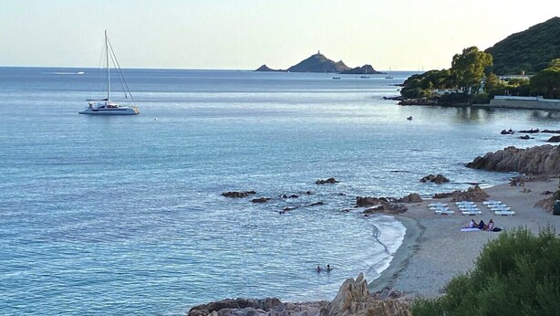 A coastal beach with rocky terrain, clear blue water, and a sailboat in the distance.