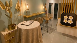 A dining area with a round table, chairs, and a vase with dried plants. There is a wooden bench with a flower painting in the background.
