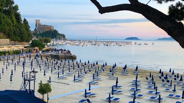 Une plage bordée de rangées de chaises longues et de parasols, une tour de surveillance pour les sauveteurs, et un château en arrière-plan.