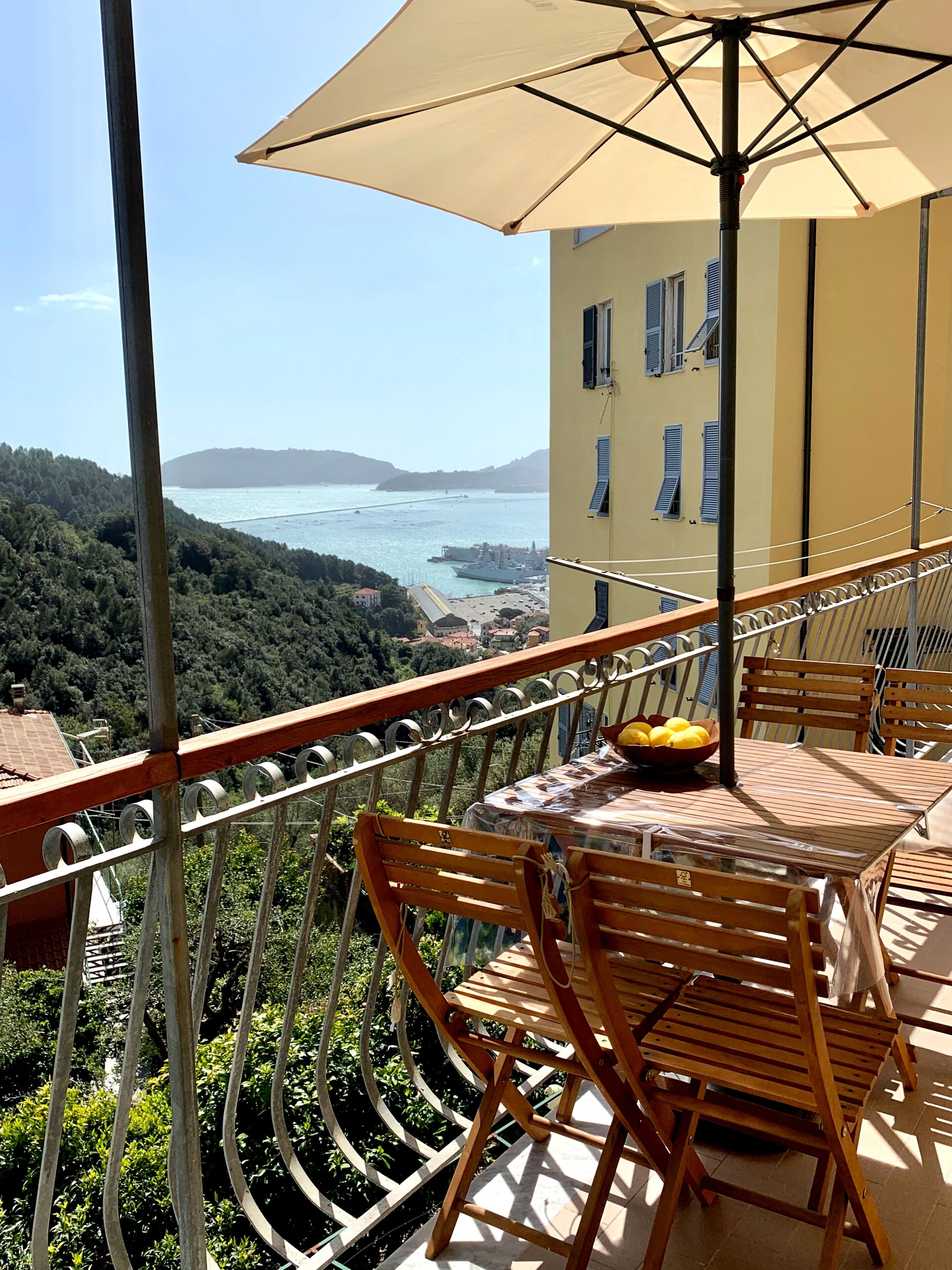 Ein Balkon mit Tisch und Stühlen, der auf eine Küstenlandschaft mit Schiff und Hügeln im Hintergrund blickt.