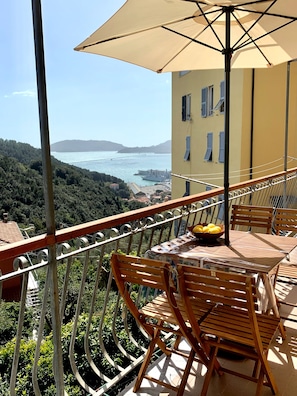 A balcony with a table and chairs, overlooking a coastal view with a ship and hills in the distance.