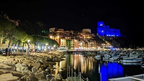 Une ville côtière nocturne, avec un château historique illuminé de bleu, une marina remplie de bateaux et des bâtiments le long du front de mer.