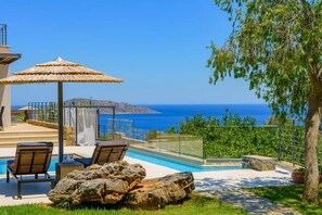 Un espace piscine doté de chaises longues et d’une pergola, offrant une vue panoramique sur la mer et les collines.