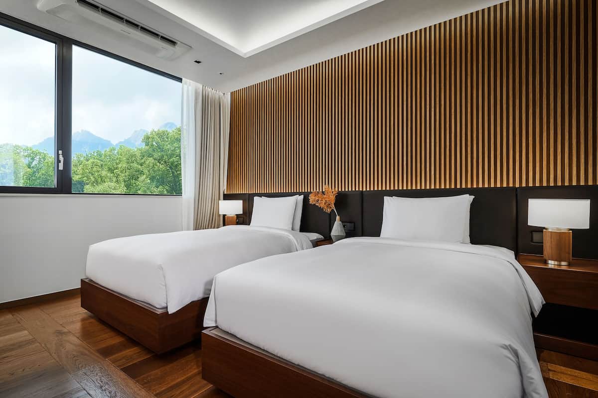 A hotel room with two beds, a wooden slat wall, a large window with a mountain view, and a nightstand with a lamp.