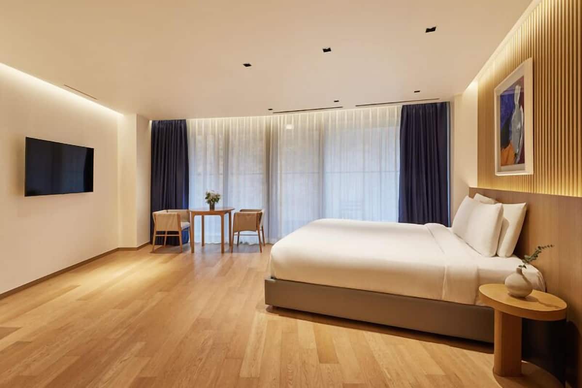 A modern hotel room with a large bed, a desk with chairs, a TV mounted on the wall, and a window with curtains.