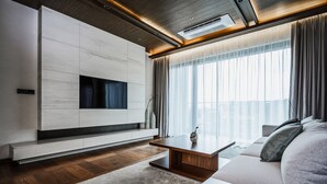 A modern living room with a flat-screen TV mounted on a white wall, a wooden coffee table, a beige sofa, and a large window with sheer curtains. - ANTO (Seoul)