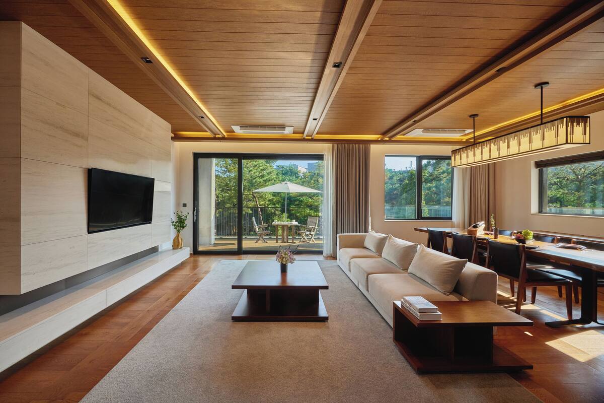 A modern living room with a flat-screen TV mounted on the wall, a wooden coffee table, a sofa, and a dining area with a table and chairs.