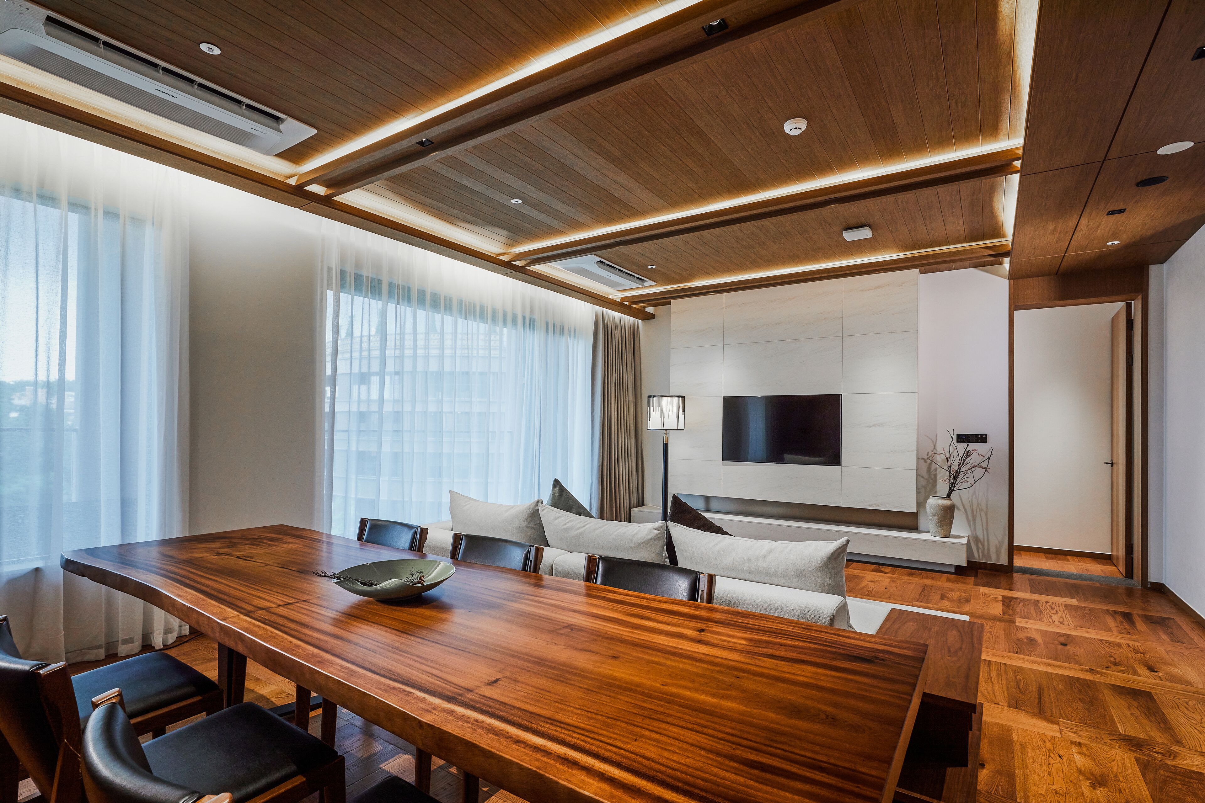 A modern conference room with a wooden table, comfortable seating, a flat-screen TV, and a large window with sheer curtains.