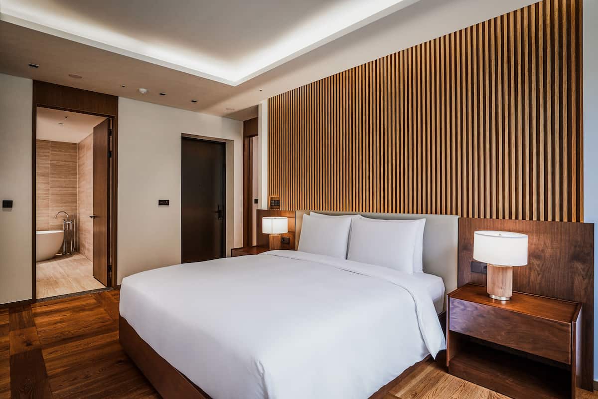 A modern hotel room with a large bed, wooden panel wall, and a nightstand with a lamp.