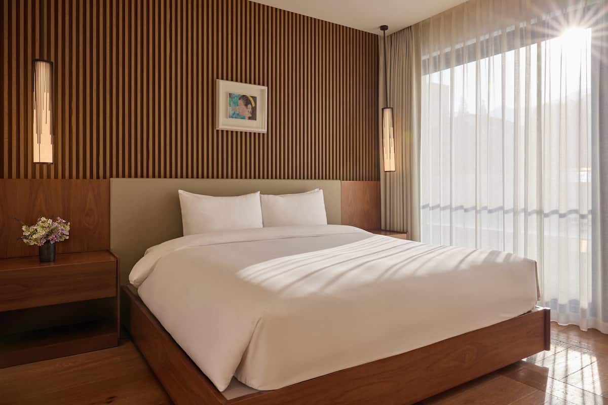 A modern hotel room with a wooden headboard, a large bed with white linens, a bedside table with a vase of flowers, and a large window with sheer curtains.