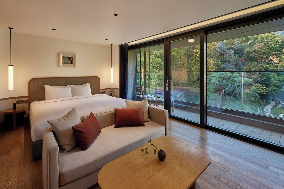 A modern hotel room with a large bed, a sofa, a wooden coffee table, and a large window offering a view of a garden.