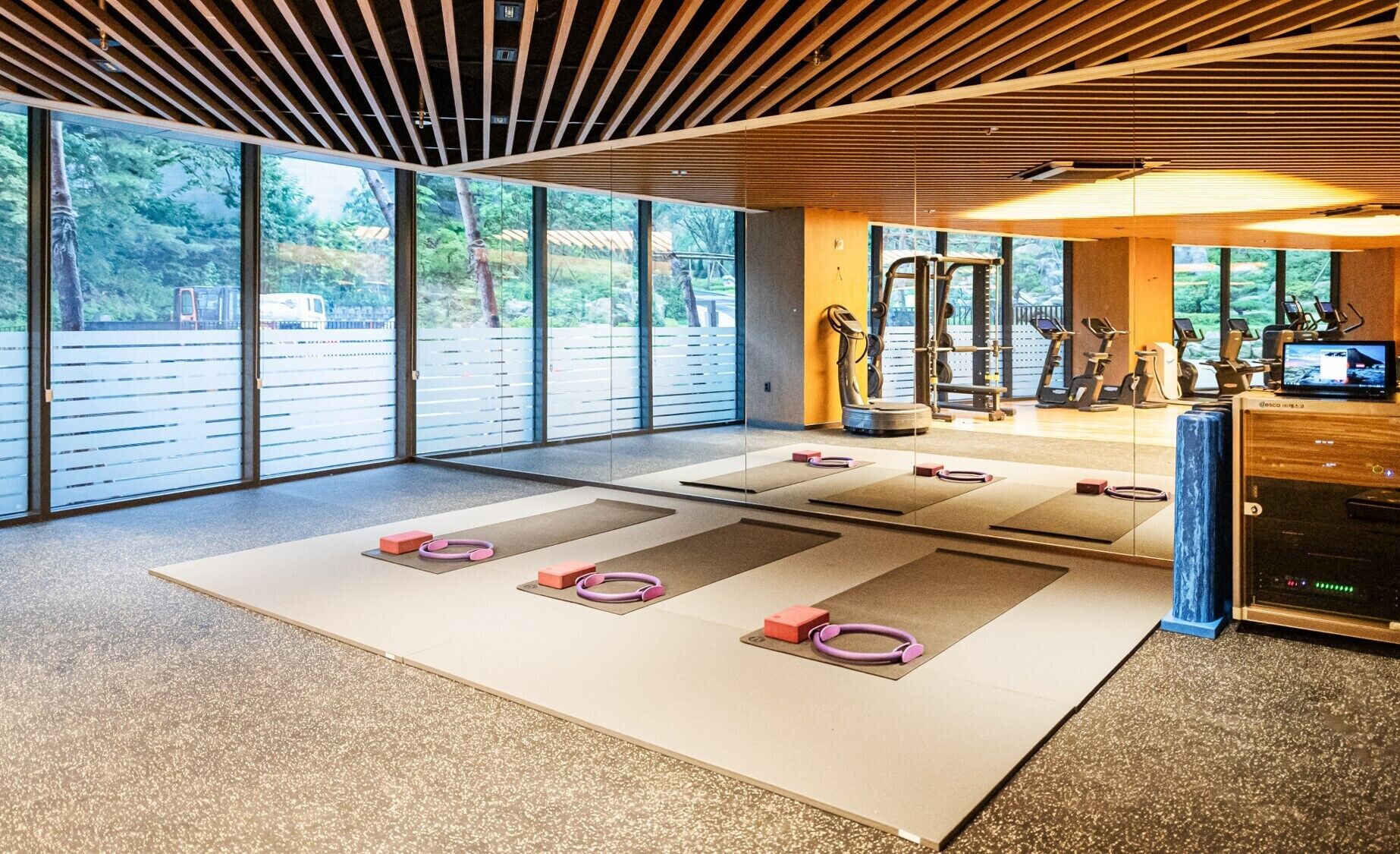 A spacious gym with exercise mats, weights, and cardio machines. Large windows offer a view of the outdoors.