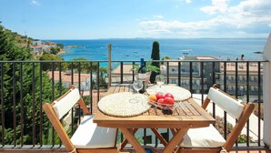 A balcony with a wooden table set for two, two white cushioned chairs, a basket of fruit, and a bottle of wine. - STA BARBARA 27 Roses (Rosas / Roses)