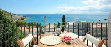 A balcony with a wooden table set for two, two white cushioned chairs, a basket of fruit, and a bottle of wine.